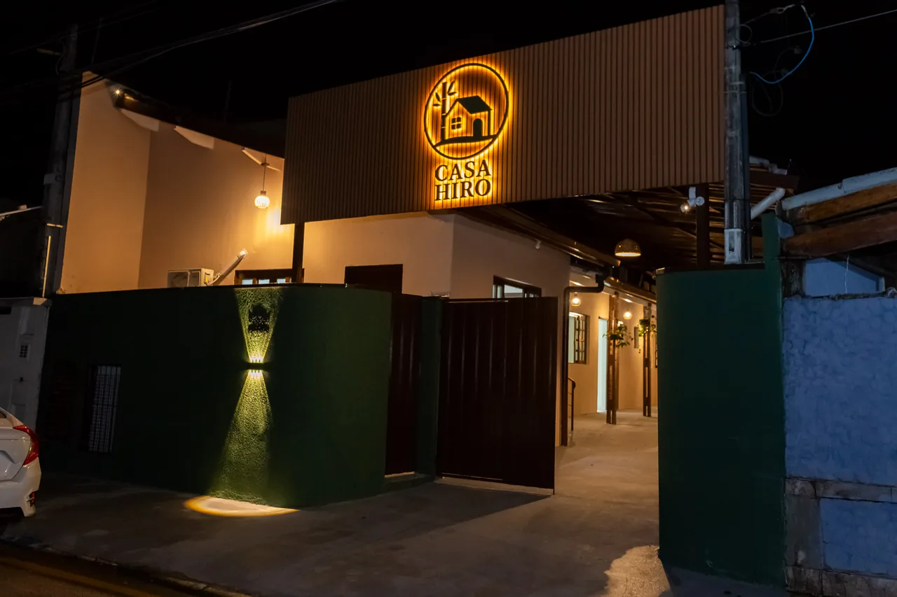 Fachada da Casa Hiro à noite, com a placa iluminada e o portão aberto